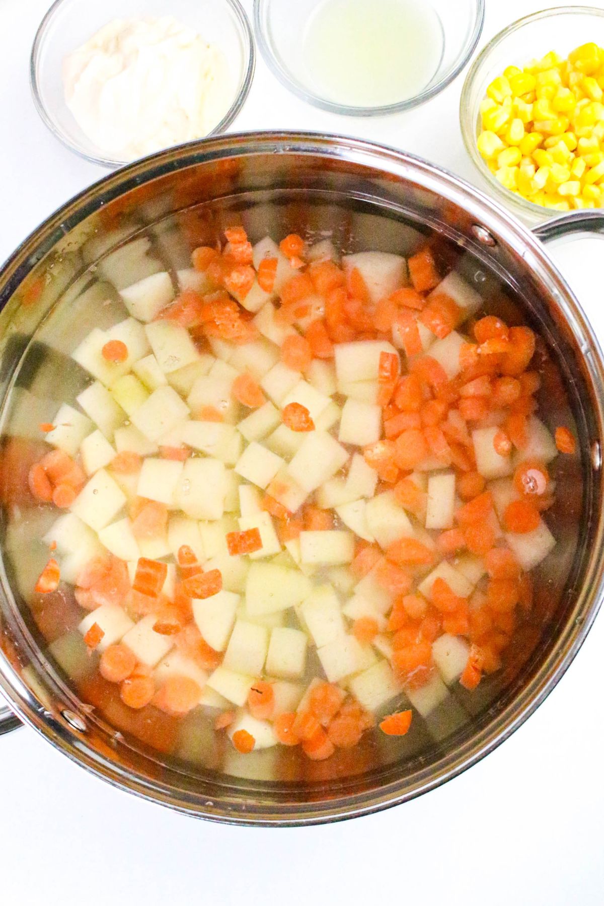A pot filled with diced potatoes and carrots in water, with small bowls of corn, mayonnaise, and a clear liquid on the side.