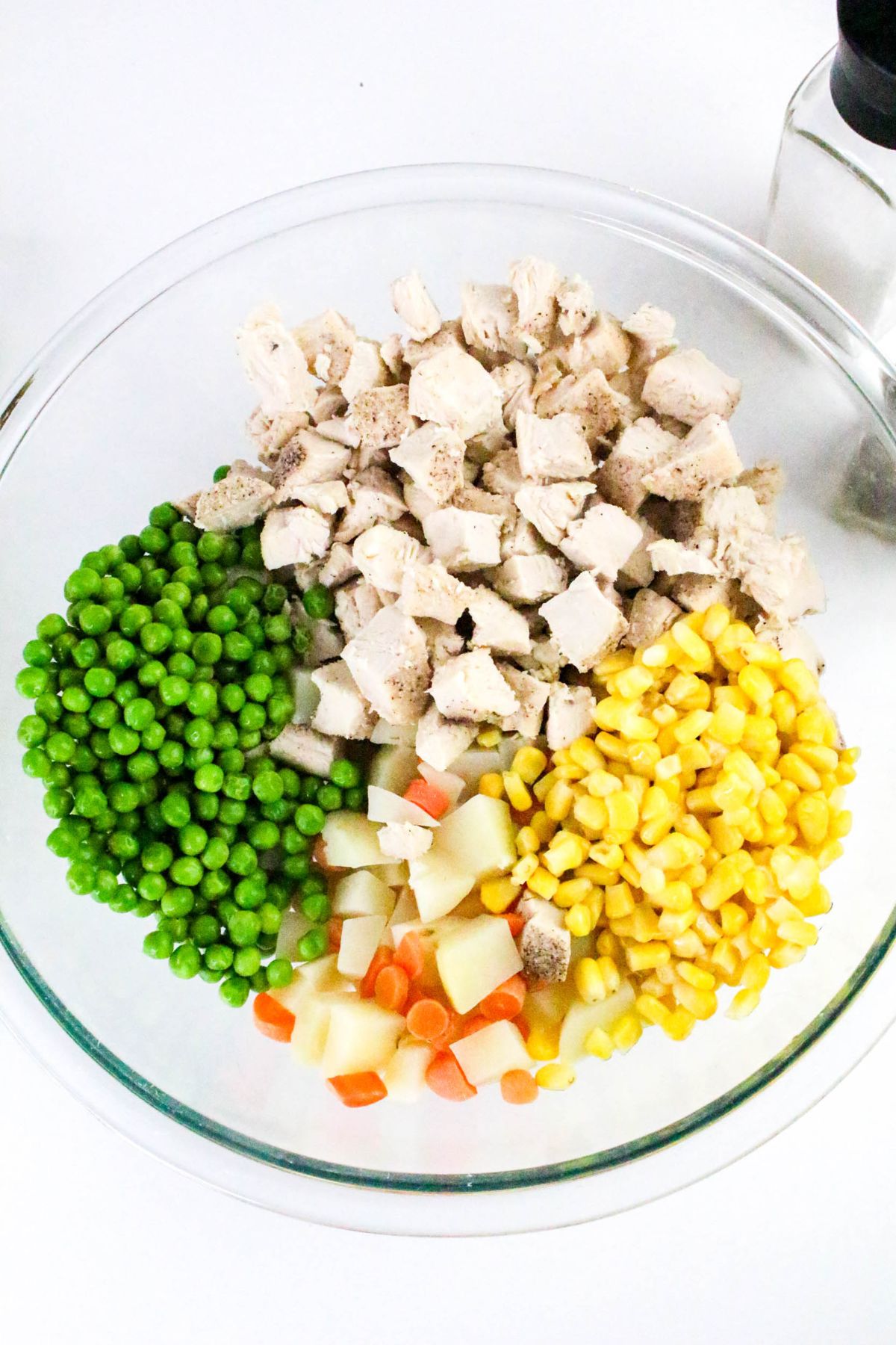 A glass bowl containing separate piles of diced cooked chicken, green peas, corn, and a mix of diced carrots and potatoes.