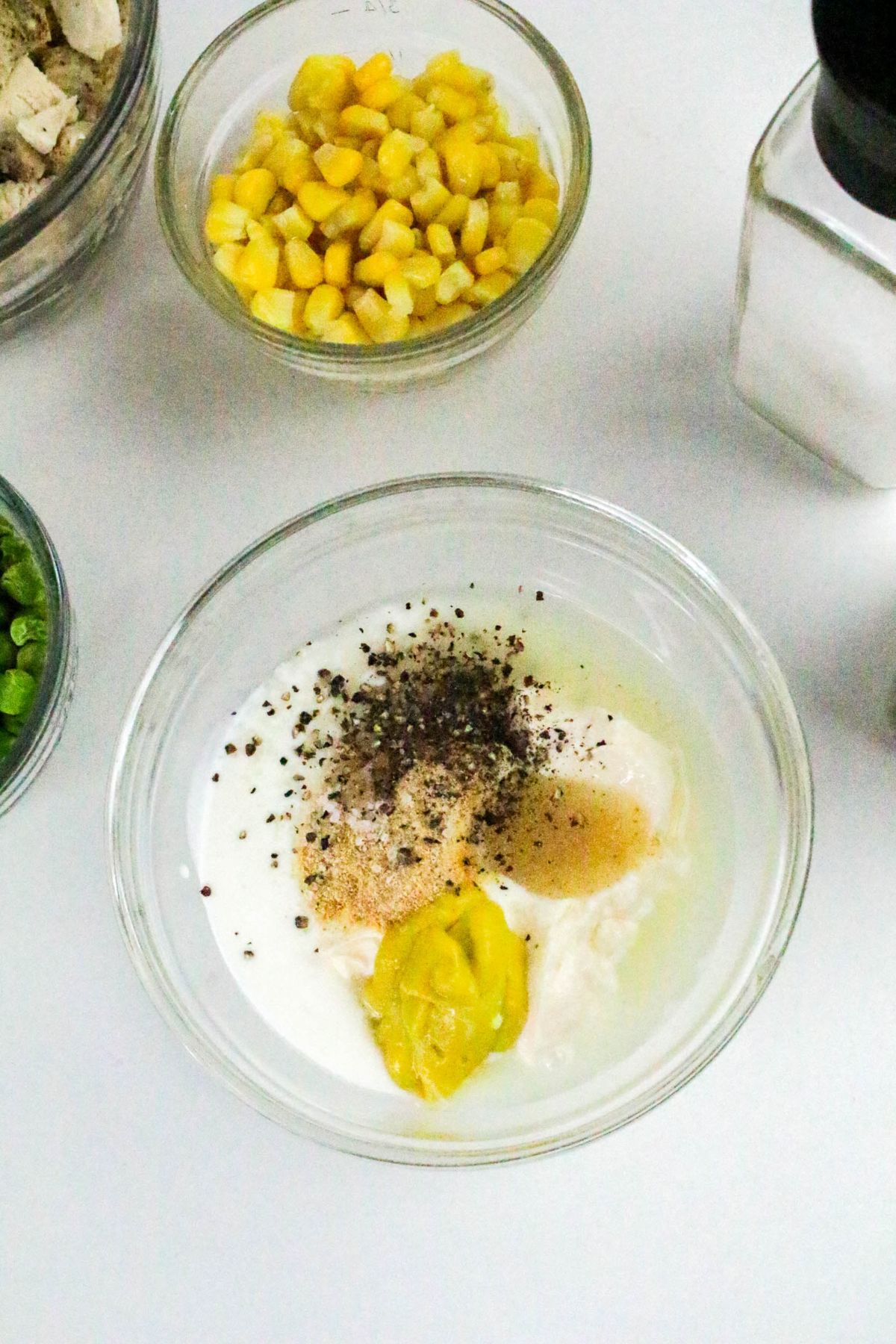 A glass bowl with yogurt, mustard, spices, and pepper, surrounded by bowls of corn, green peas, and a pepper grinder on a white surface.