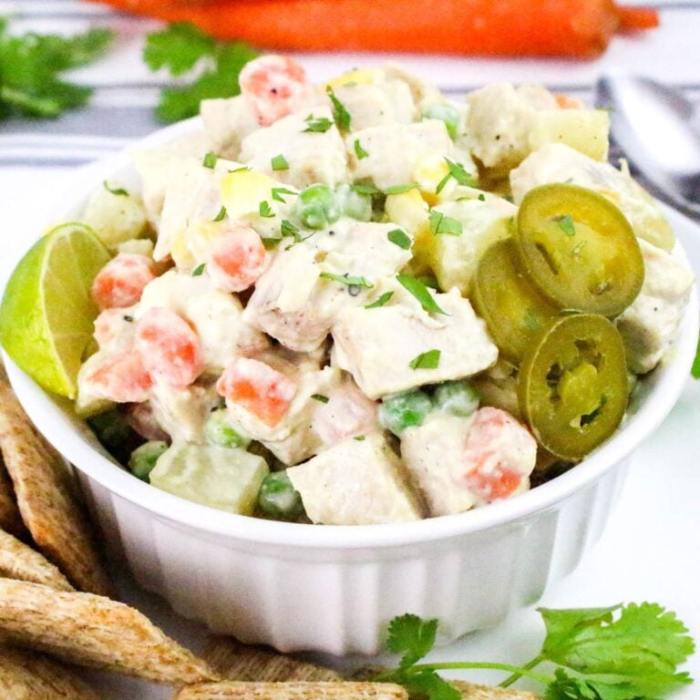A bowl of creamy chicken salad with diced carrots, peas, potatoes, lime wedge, sliced jalapeños, and herbs, served with bread strips.