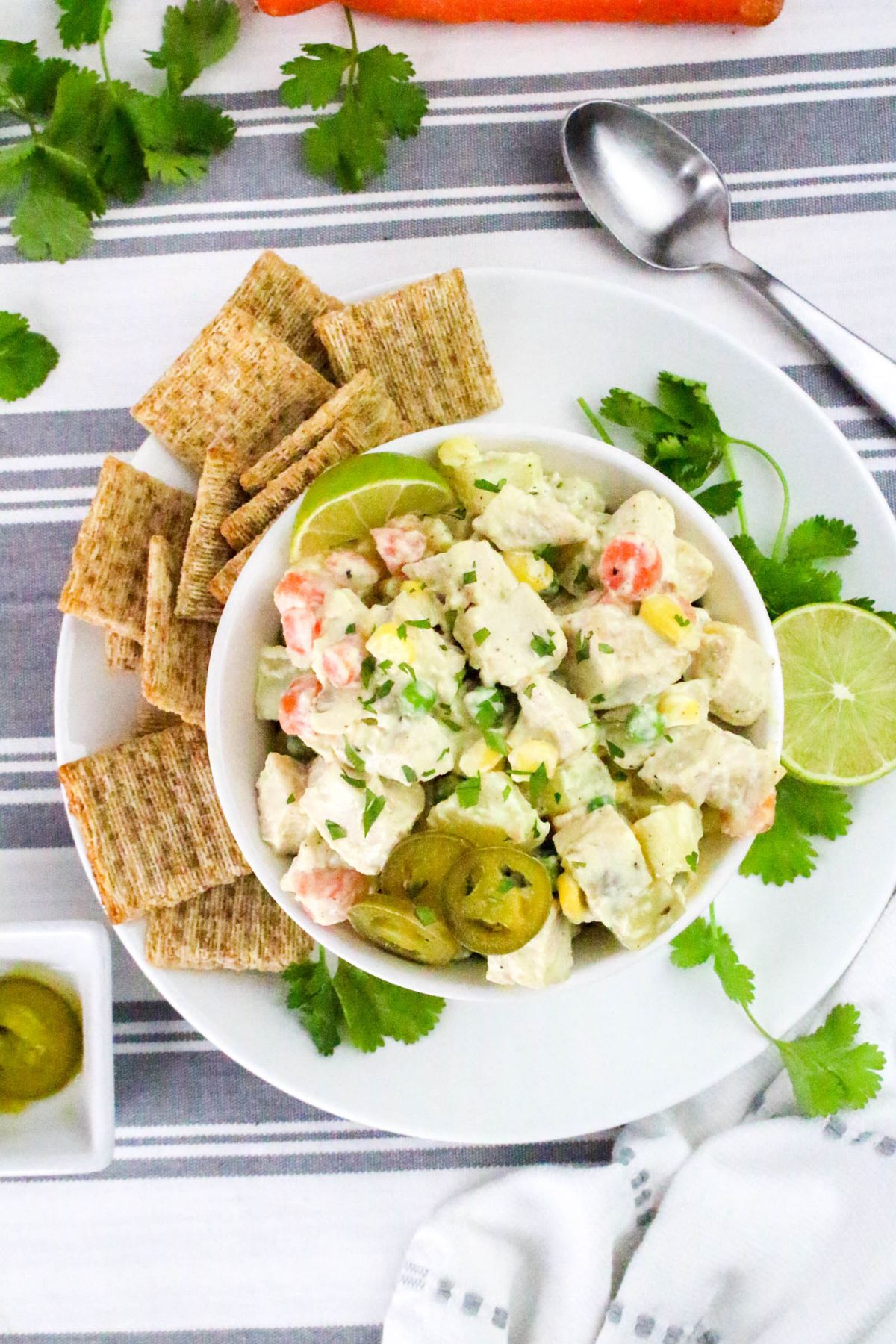A bowl of potato salad with vegetables, garnished with jalapeño slices and cilantro, served with lime wedges and a side of woven crackers on a white plate.
