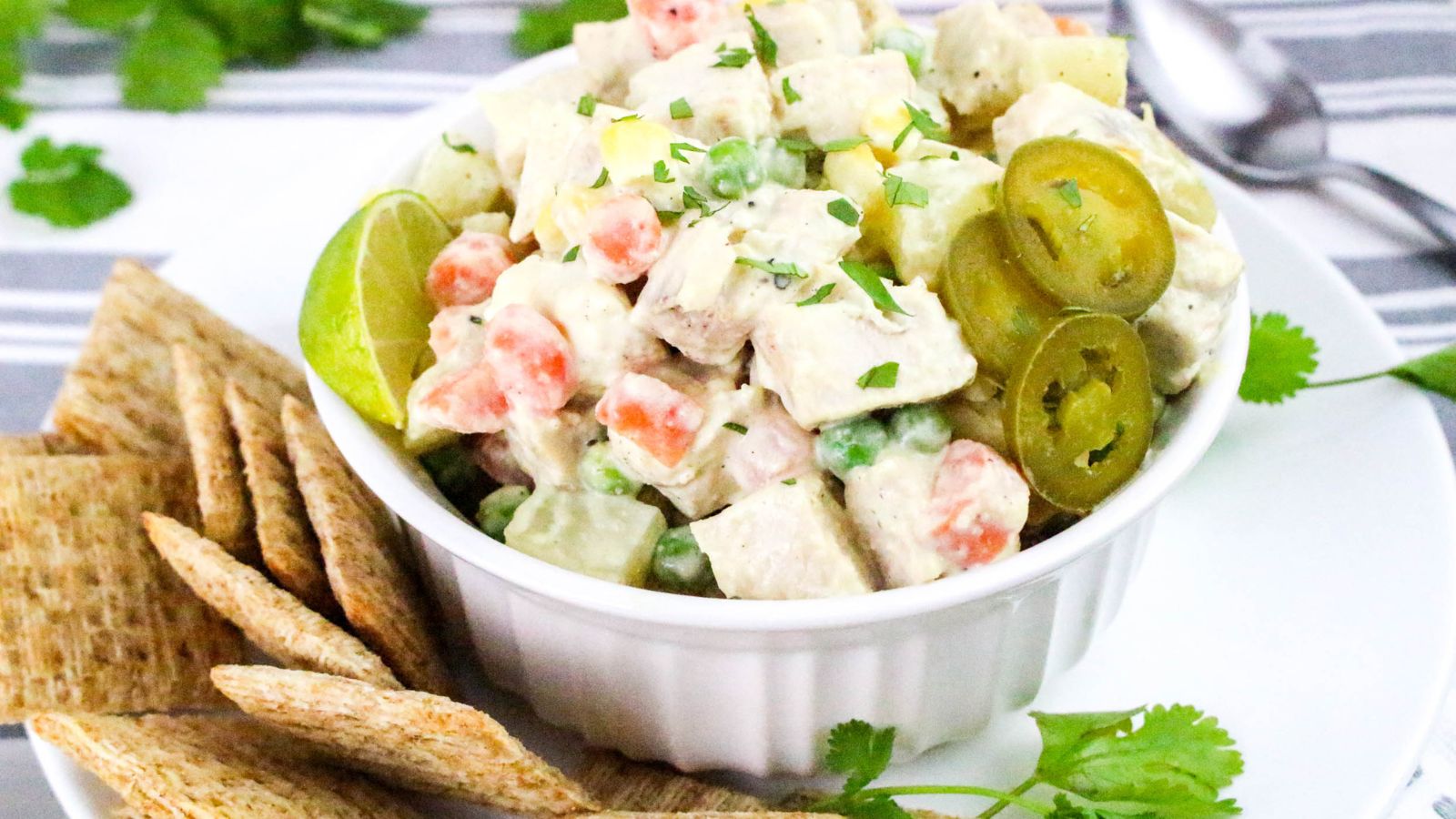 A bowl of chicken salad with diced vegetables, lime wedges, sliced jalapeños, and fresh herbs, served with crackers on a white plate.