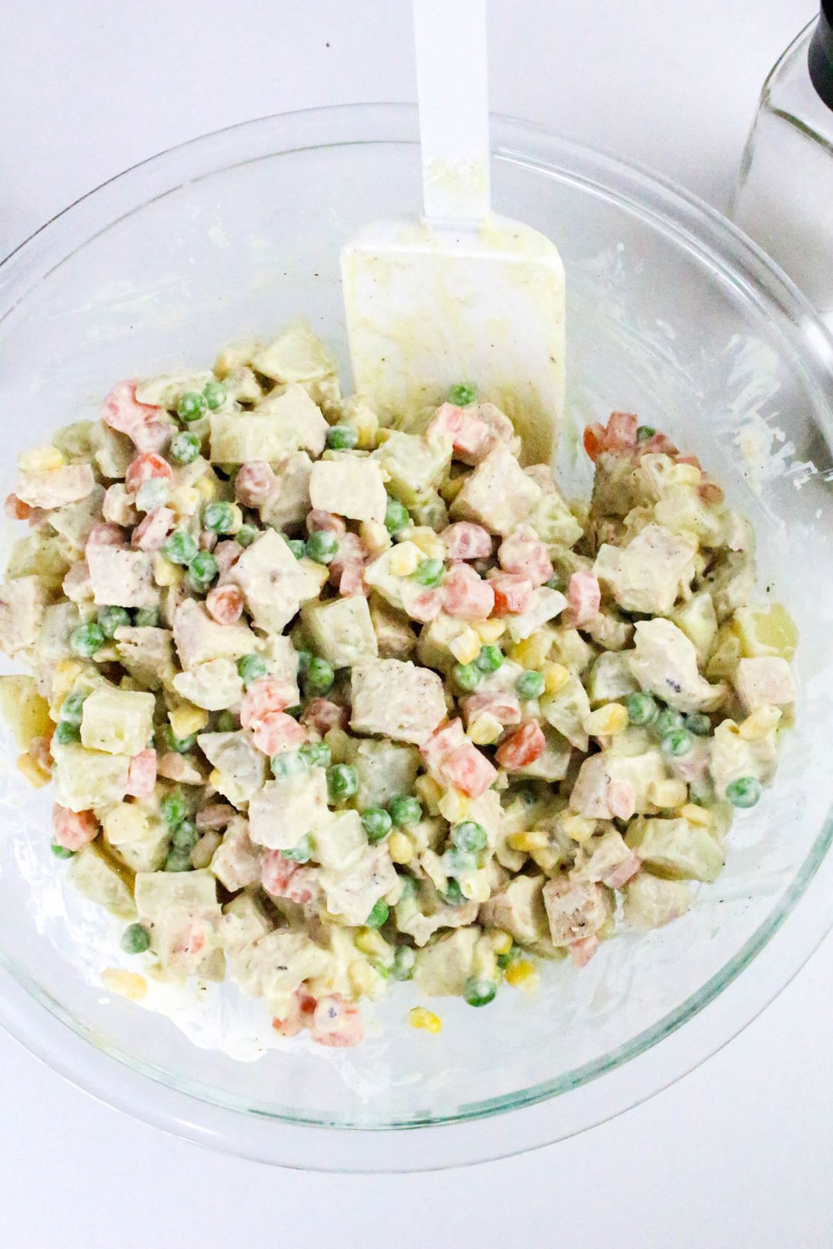 A glass bowl of mixed potato salad with diced potatoes, peas, carrots, and corn, coated in a creamy dressing, with a white spatula resting inside.
