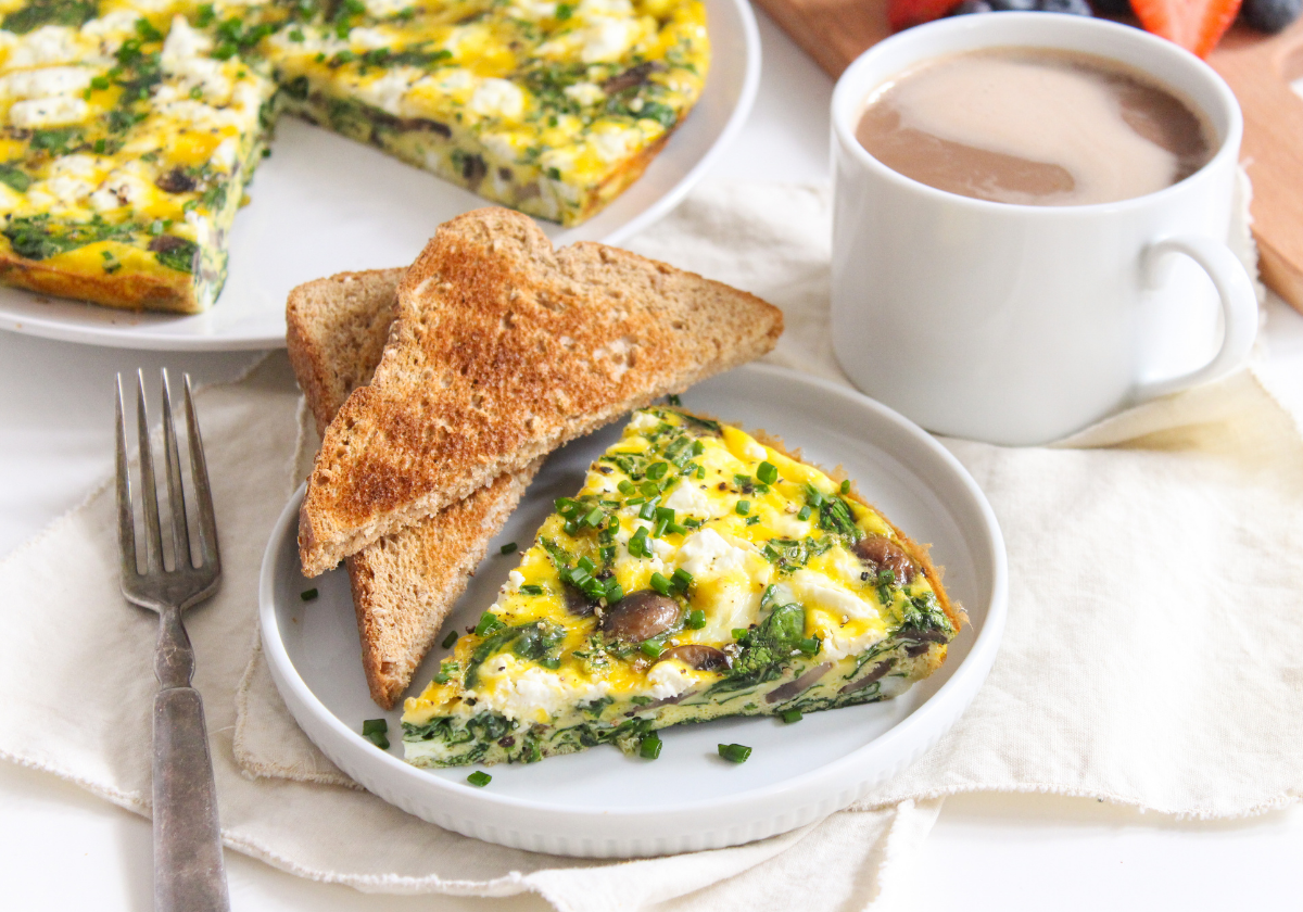A slice of mushroom frittata on a white plate beside a slice of toast, with a mug of coffee in the background.