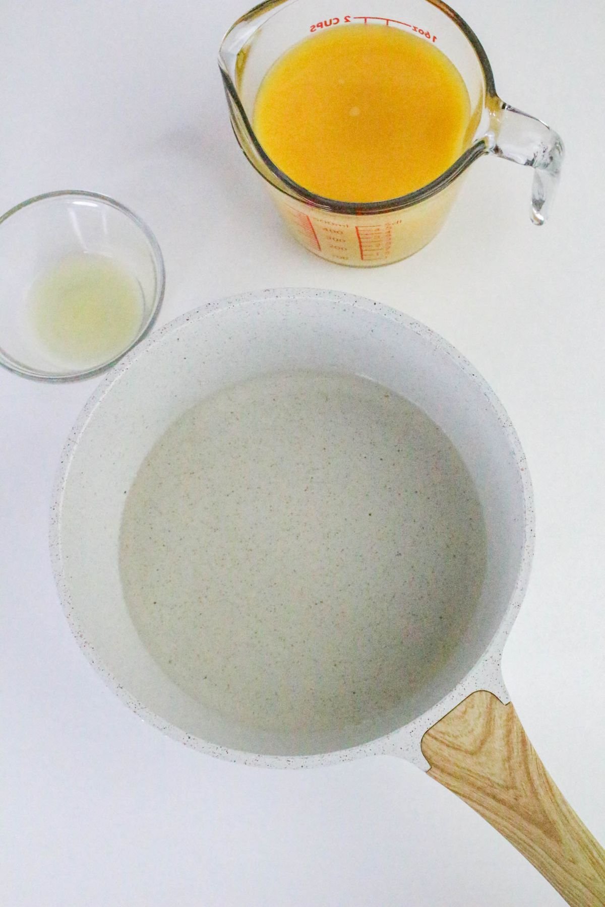A white saucepan with clear liquid, a small glass bowl with more clear liquid, and a measuring cup with orange liquid are arranged on a white surface.