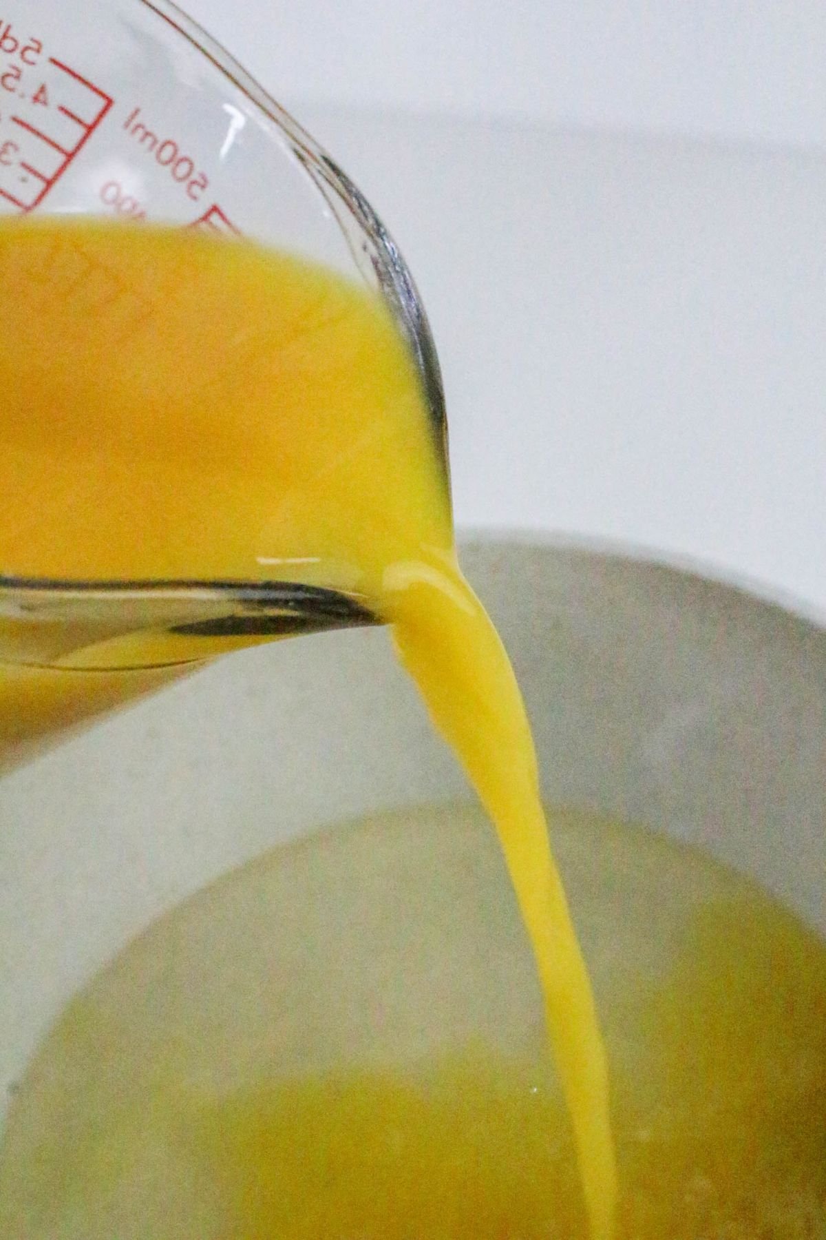 Orange liquid is being poured from a measuring cup into a pot containing a clear yellowish liquid.