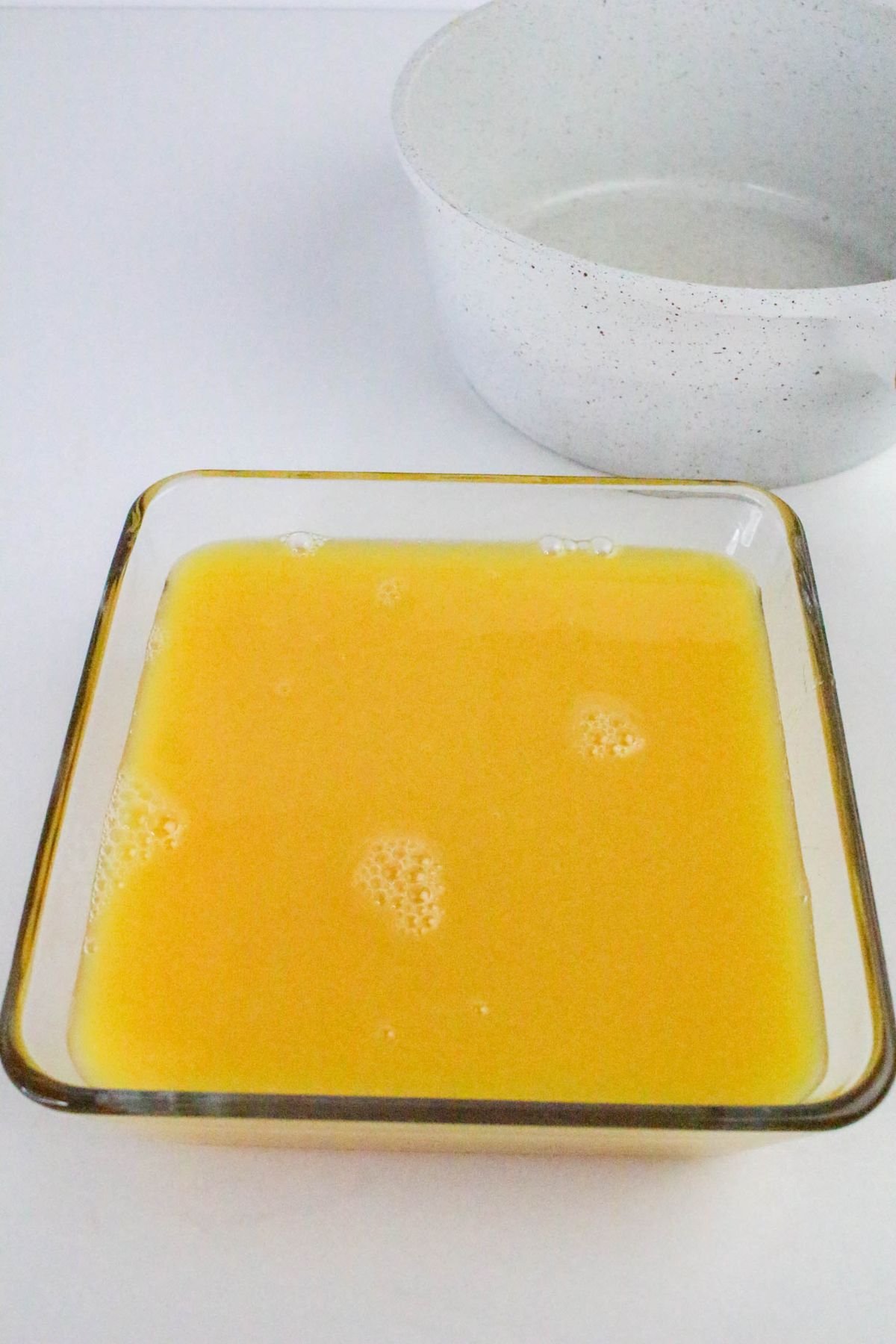 A square glass dish filled with orange liquid sits on a white surface near an empty white bowl.