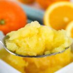 A close-up of a spoonful of orange granita with fresh oranges and orange slices in the blurred background.