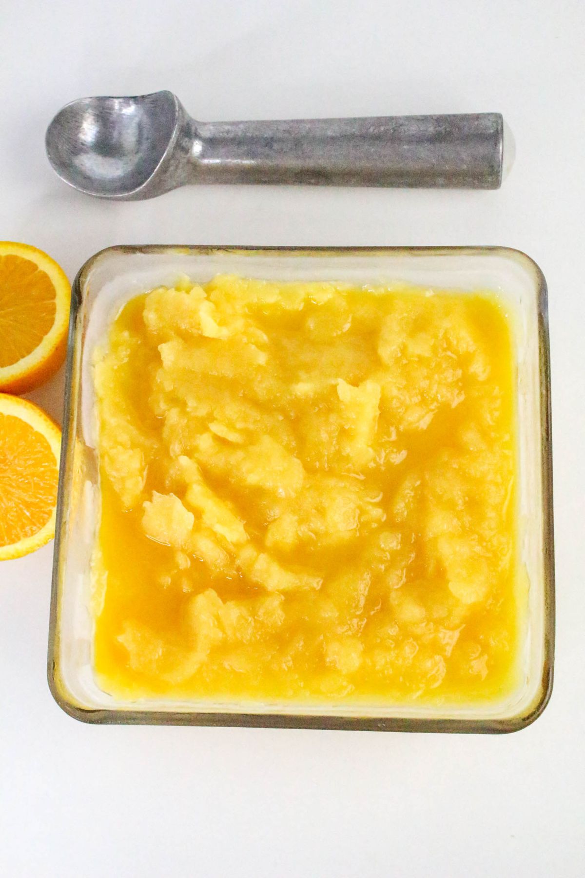 A glass dish filled with orange sorbet sits on a white surface next to an ice cream scoop and two orange halves.