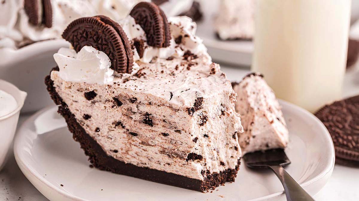 A slice of cookies and cream pie with whipped topping and chocolate cookie halves sits on a white plate with a fork.