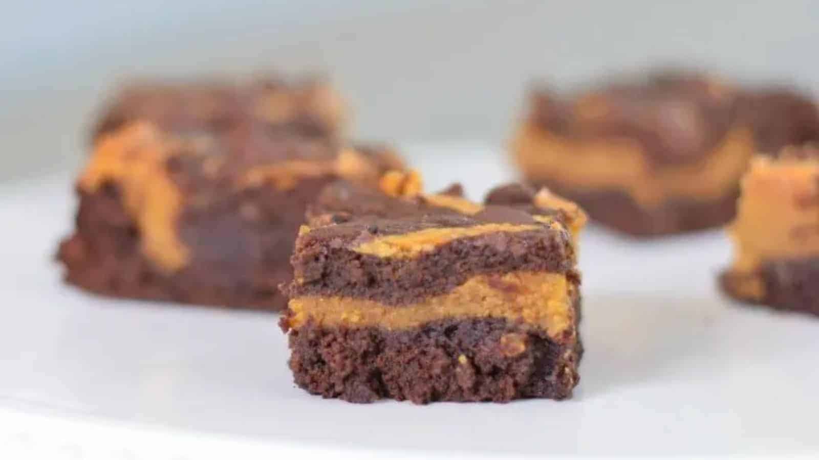 A close-up of a chocolate and peanut butter layered brownie on a white surface, with more brownies blurred in the background.