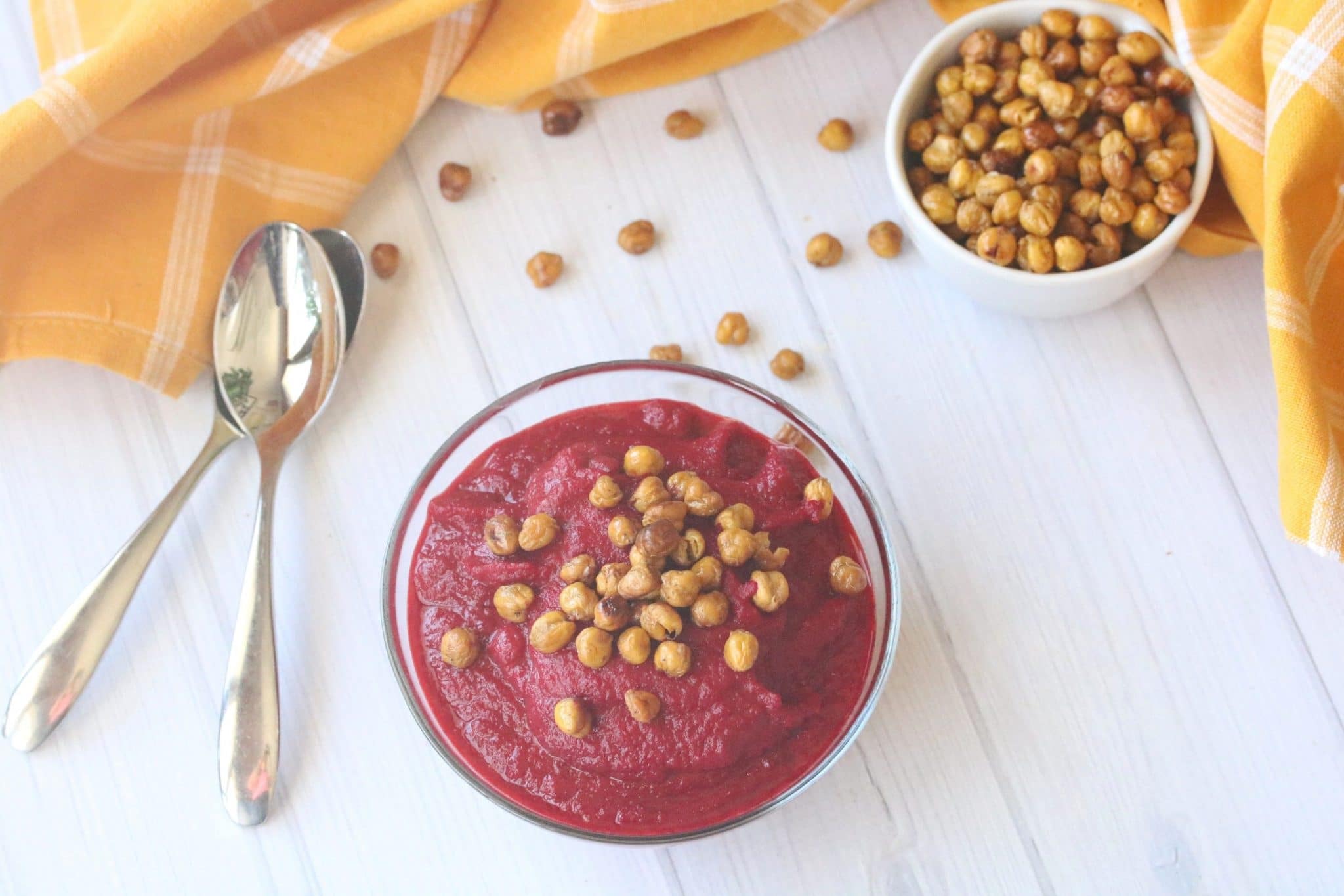 two glass bowls filled with healthy beet soup and topped with chickpeas. Two spoons beside the first bowl and a yellow napkin.