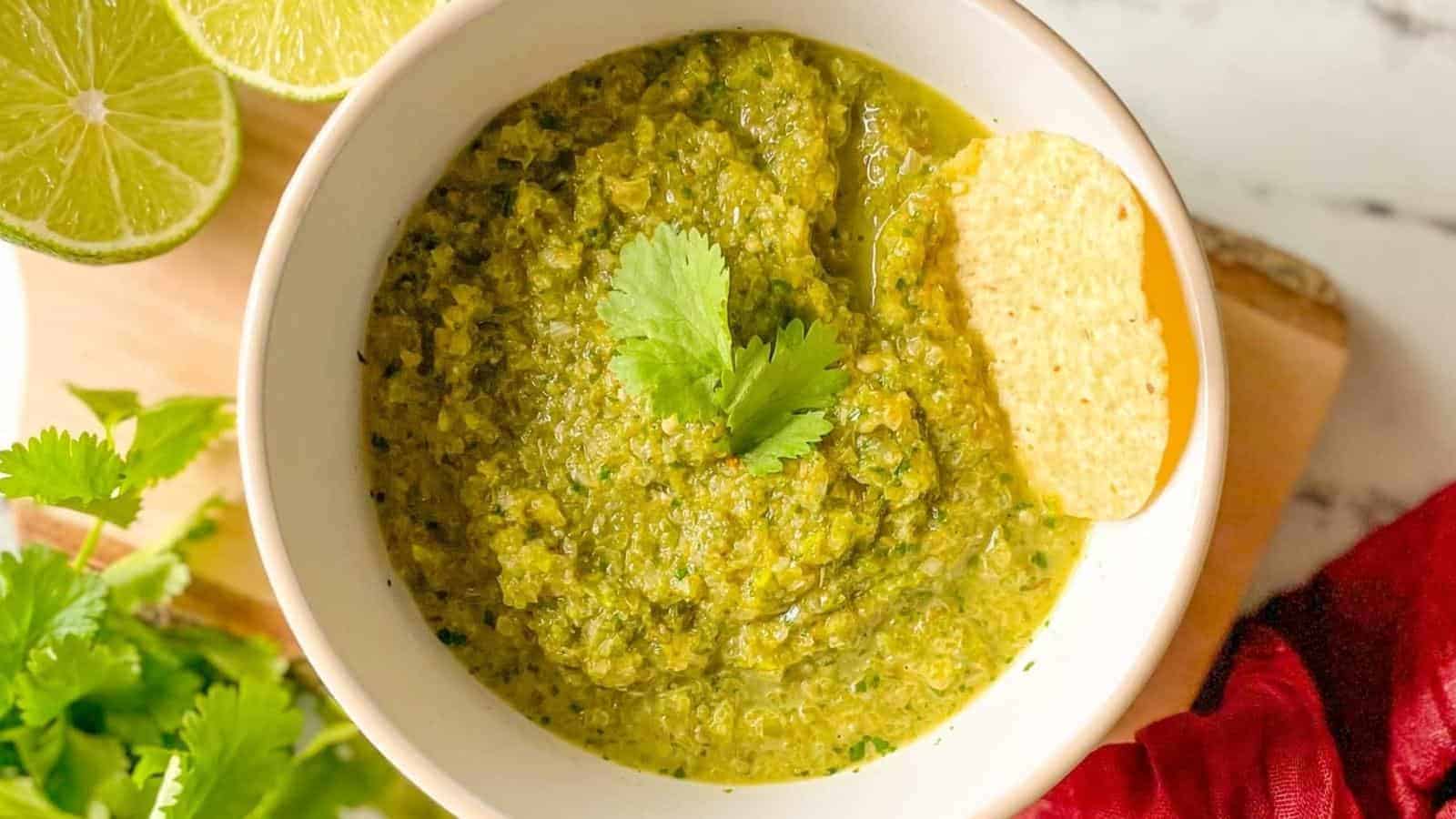 A bowl of serrano salsa is shown with limes, chips, cilantro, and a red linen.