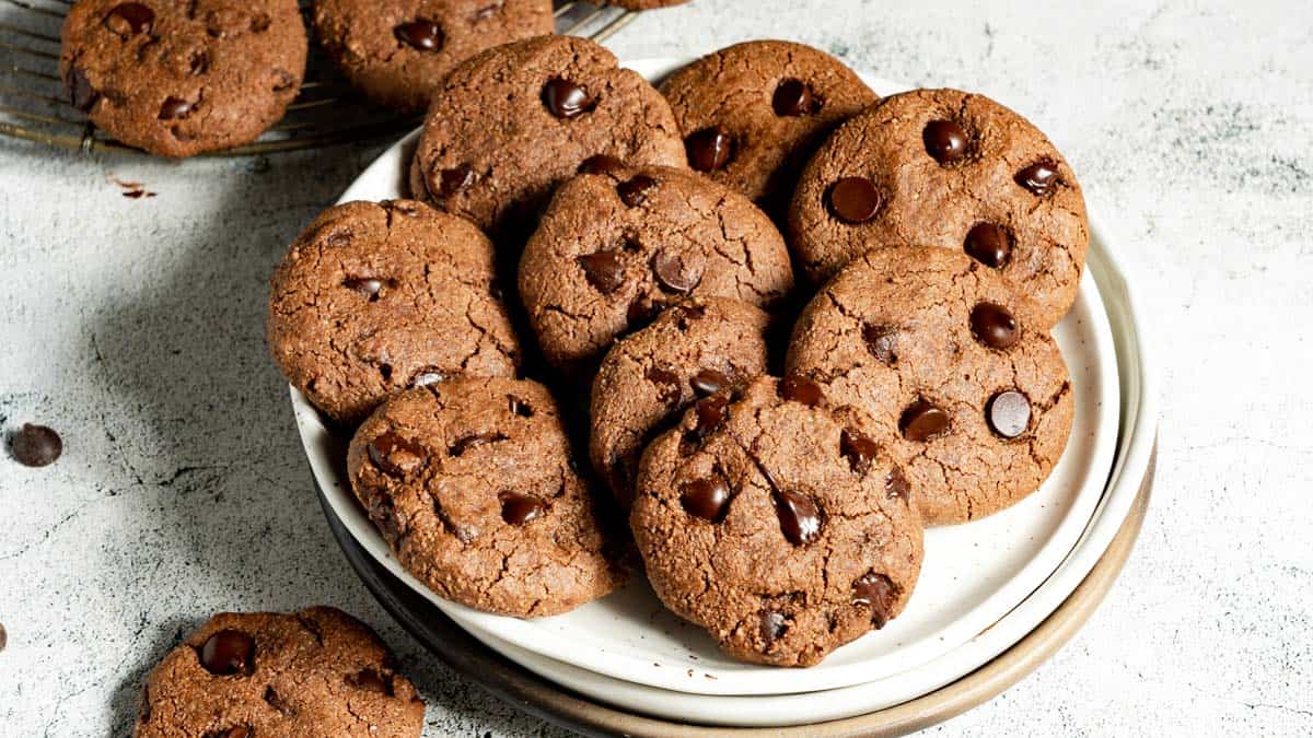 A plate of chocolate chip cookies is arranged on a light surface, with additional cookies partially visible in the background.