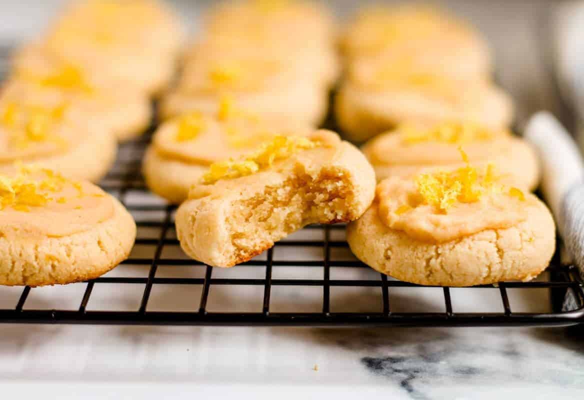 A batch of round cookies with light icing and lemon zest sits on a cooling rack; one cookie in front has a bite taken out of it.