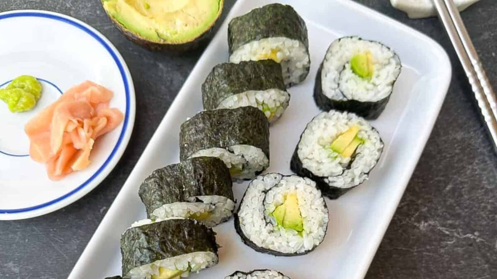 A plate of sushi rolls with avocado filling on a rectangular dish. A small plate with ginger and wasabi, and a halved avocado are beside it.