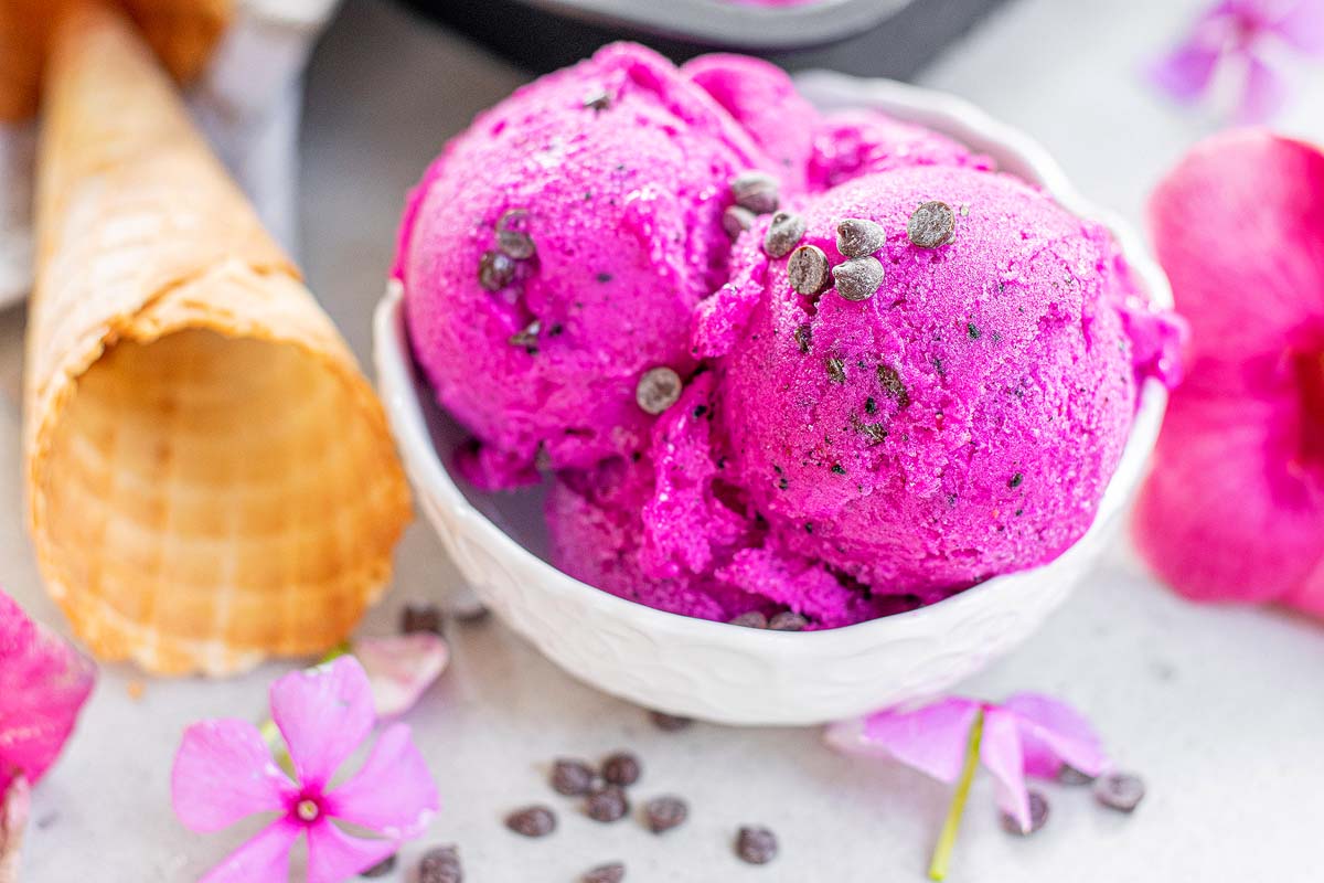 A white bowl with two scoops of bright pink ice cream topped with chocolate chips, next to an empty waffle cone and pink flowers.