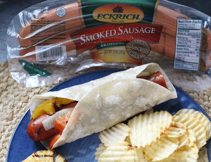 A smoked sausage in a tortilla with grilled vegetables, served on a blue plate with potato chips. The sausage package is visible in the background.