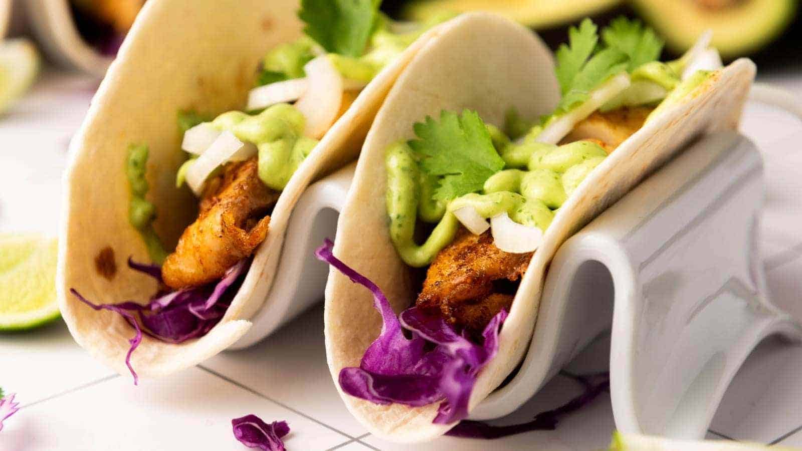 Two fish tacos filled with grilled fish, shredded purple cabbage, chopped onions, cilantro, and drizzled with a green sauce.