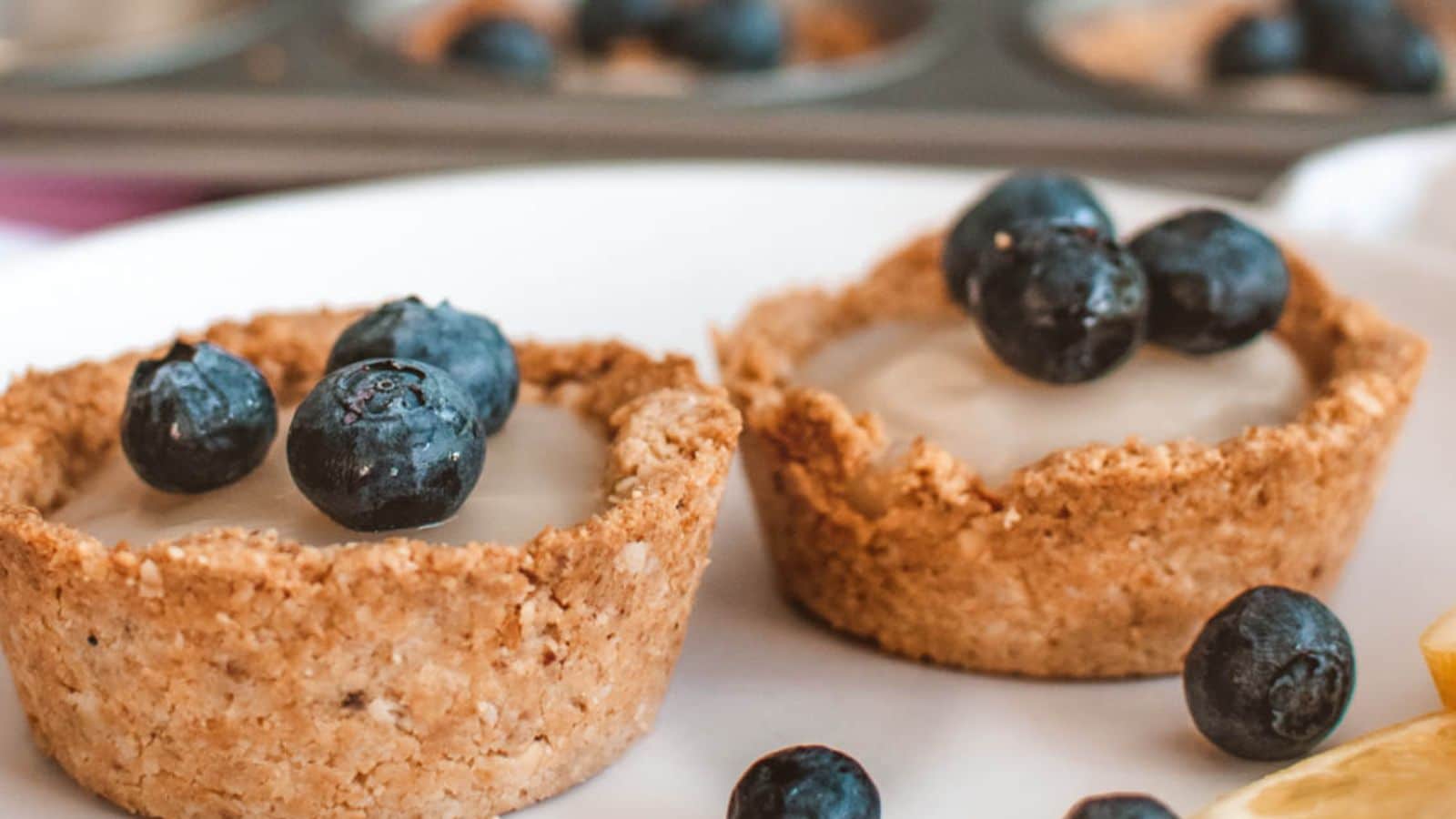 Two small tartlets with creamy filling are topped with fresh blueberries on a white plate, with additional blueberries and part of a lemon slice nearby.