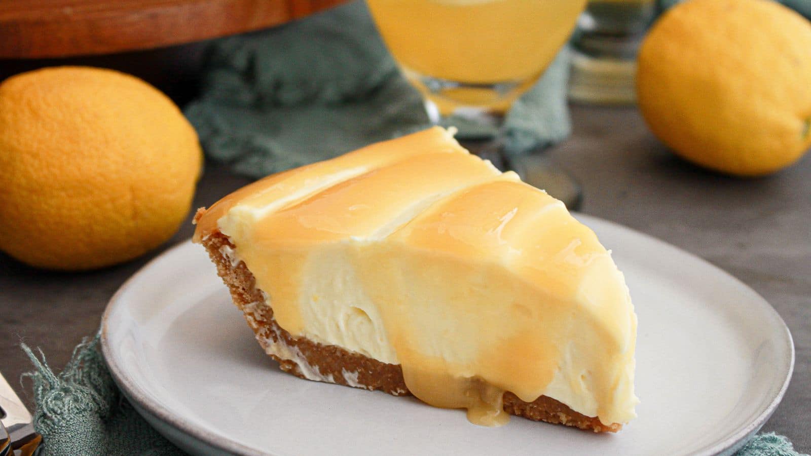 A slice of cheesecake with a graham cracker crust and lemon glaze on a plate, with whole lemons and a glass of lemon drink in the background.