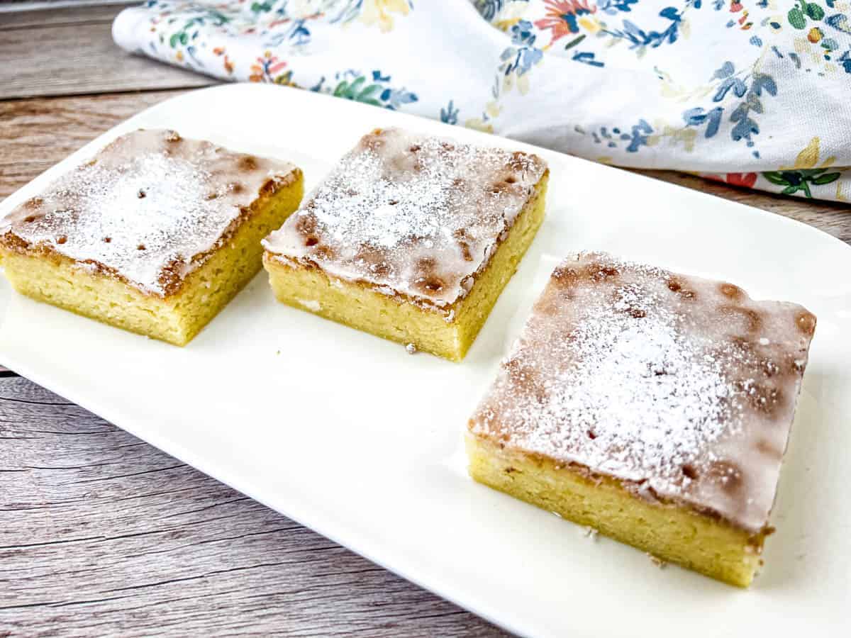 Three square pieces of glazed yellow cake with powdered sugar on top are arranged on a white rectangular plate.