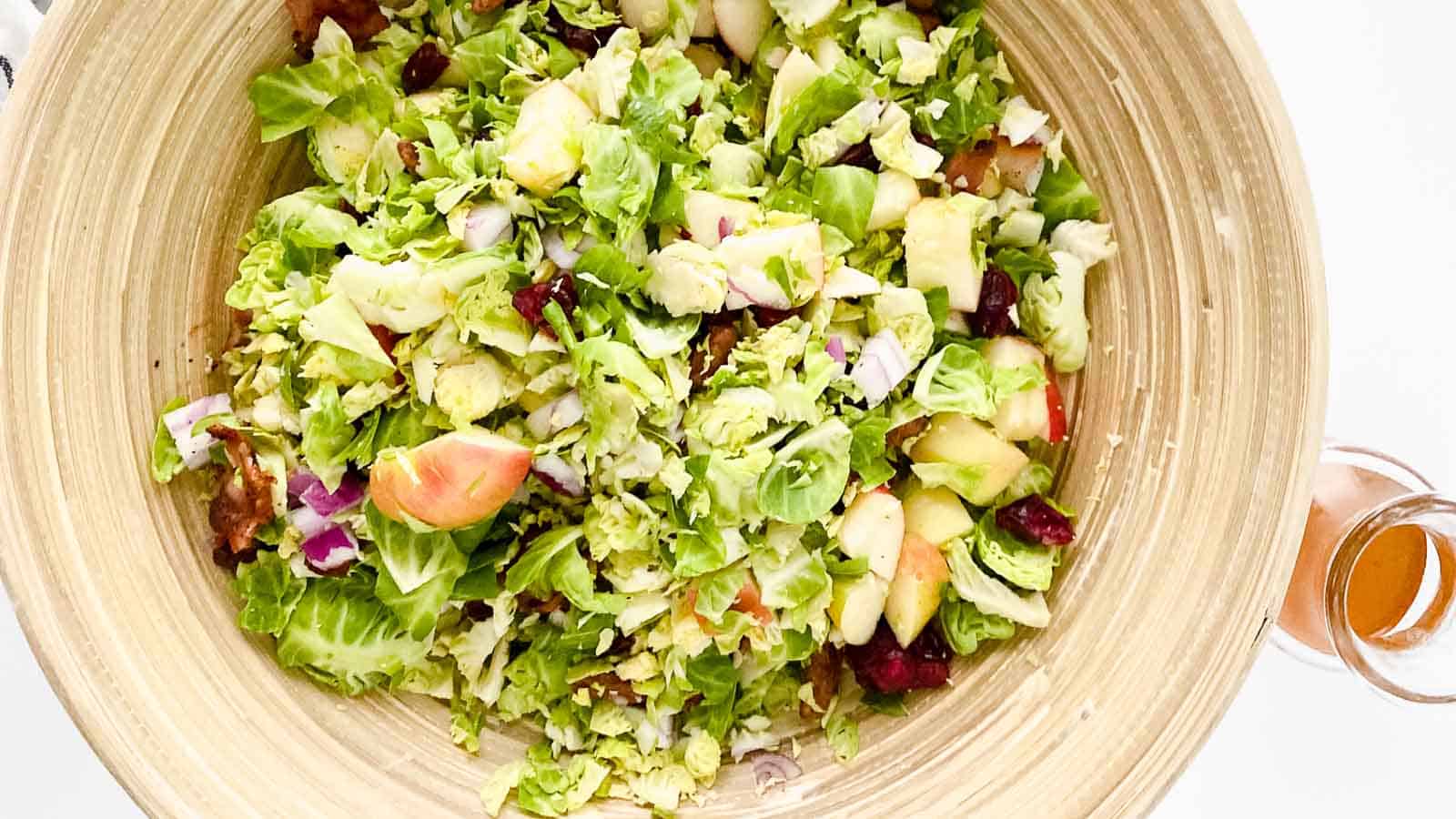 A wooden bowl filled with chopped salad ingredients including Brussels sprouts, apples, red onions, dried cranberries, and walnuts. A small glass of dressing sits nearby.