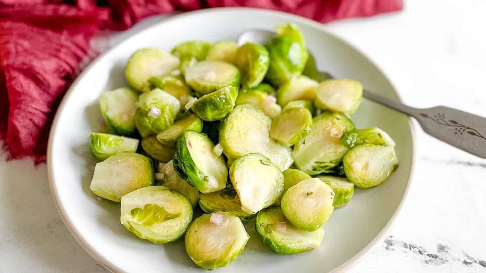 A plate of halved, cooked Brussels sprouts with small diced onions, served with a fork on a white surface.
