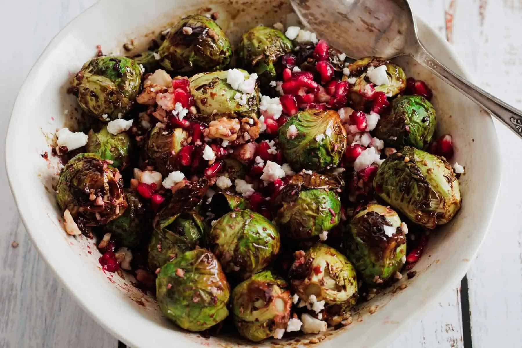 A bowl of roasted Brussels sprouts topped with feta cheese, pomegranate seeds, and chopped walnuts, with a spoon on the side.