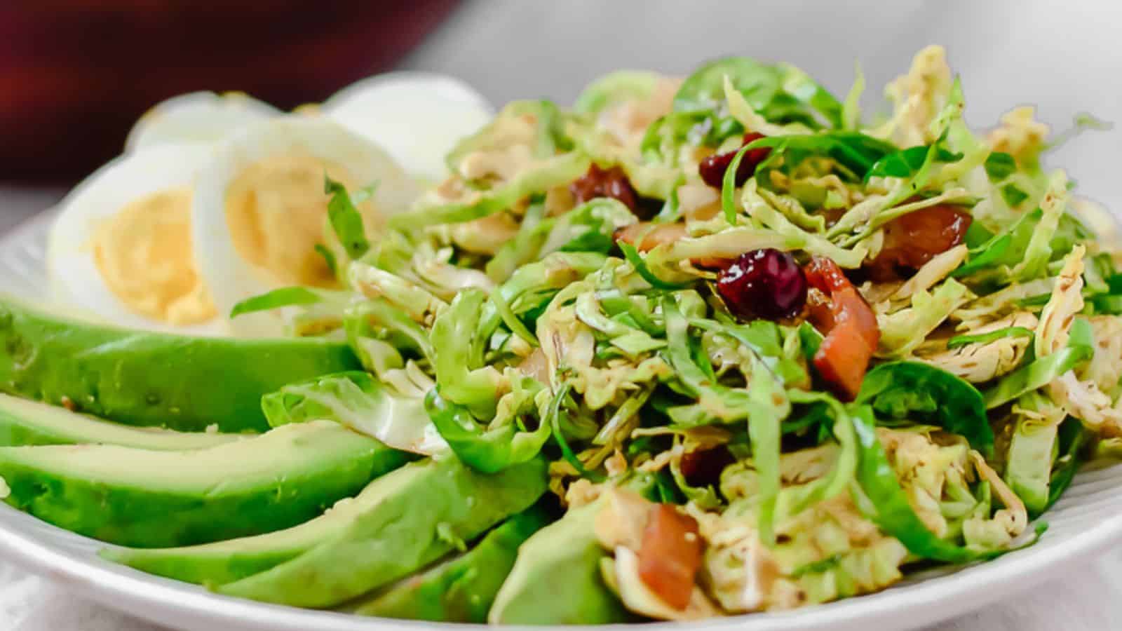 A plate with sliced avocado, halved boiled eggs, and a shredded Brussels sprout salad with dried cranberries and chopped nuts.