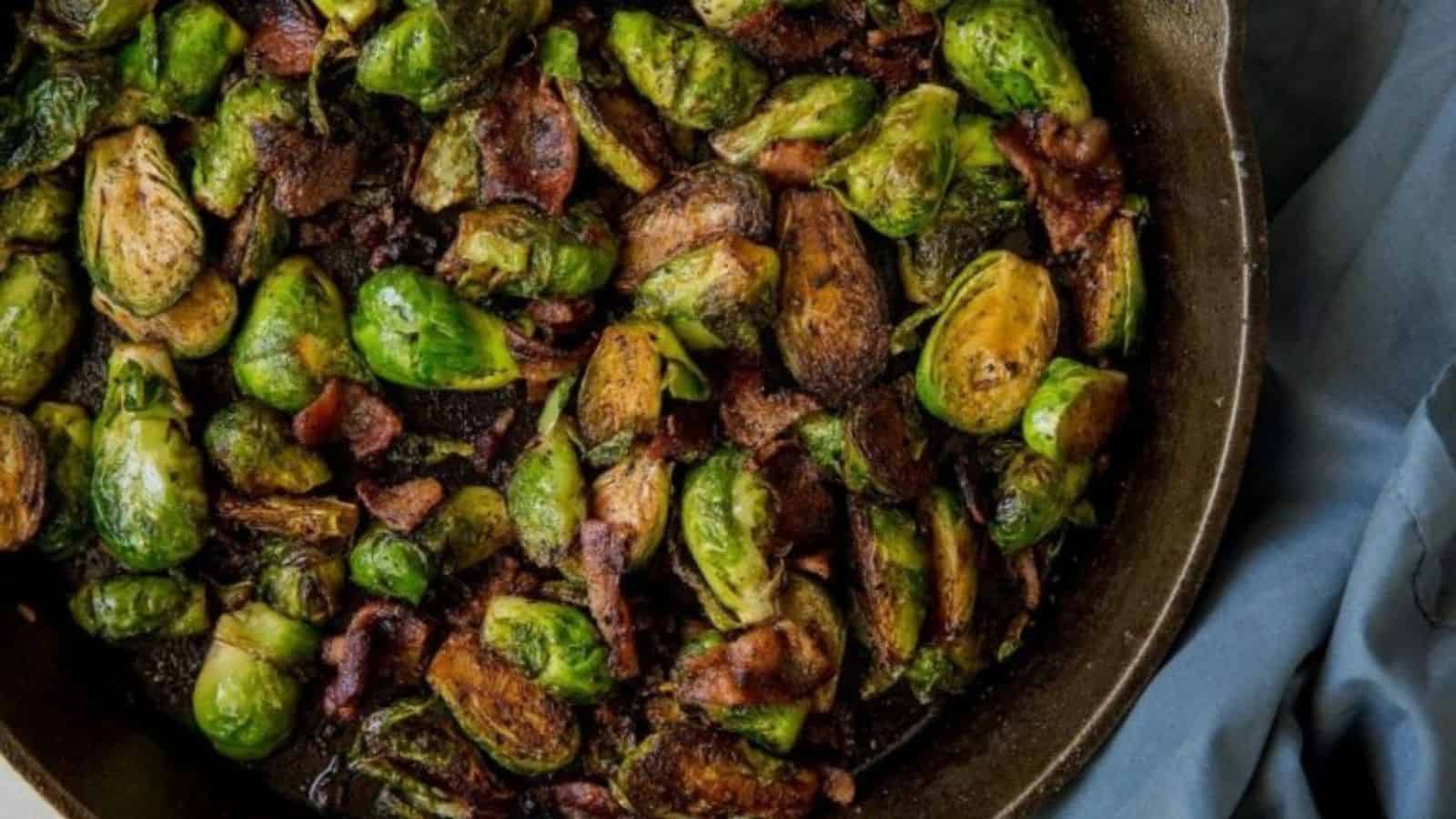 Brussels sprouts and pieces of bacon are sautéed together in a dark skillet, with slight charring visible on some of the sprouts.