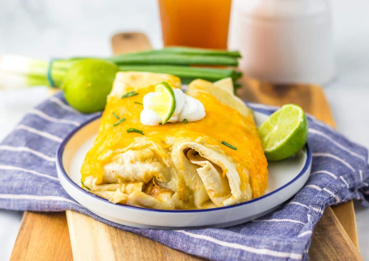 A plate with two cheesy enchiladas topped with sour cream and lime slices, garnished with chives, sits on a striped cloth with limes, scallions, and a drink in the background.