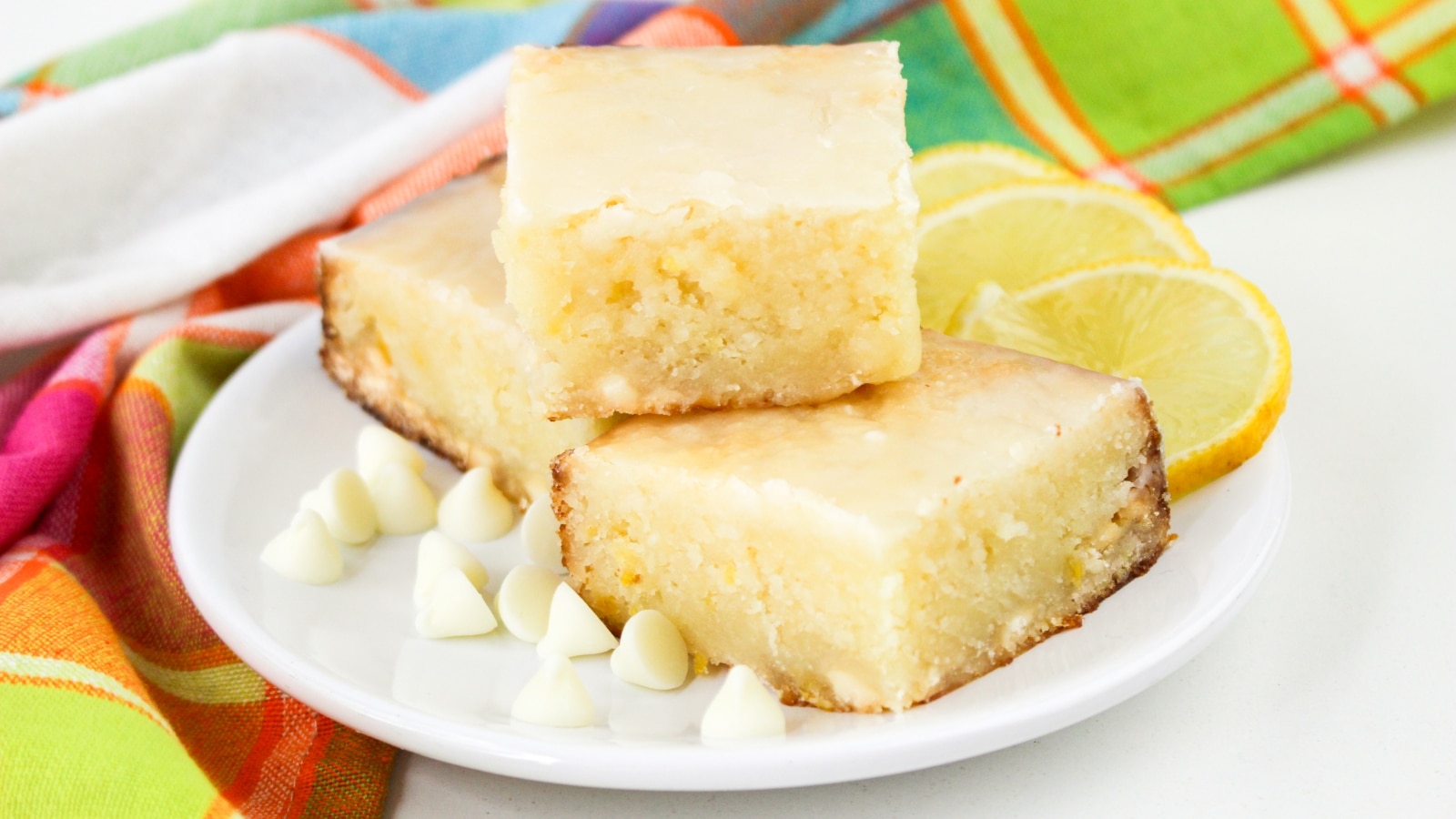 Three lemon blondies are stacked on a white plate with white chocolate chips and lemon slices, set beside a colorful cloth.