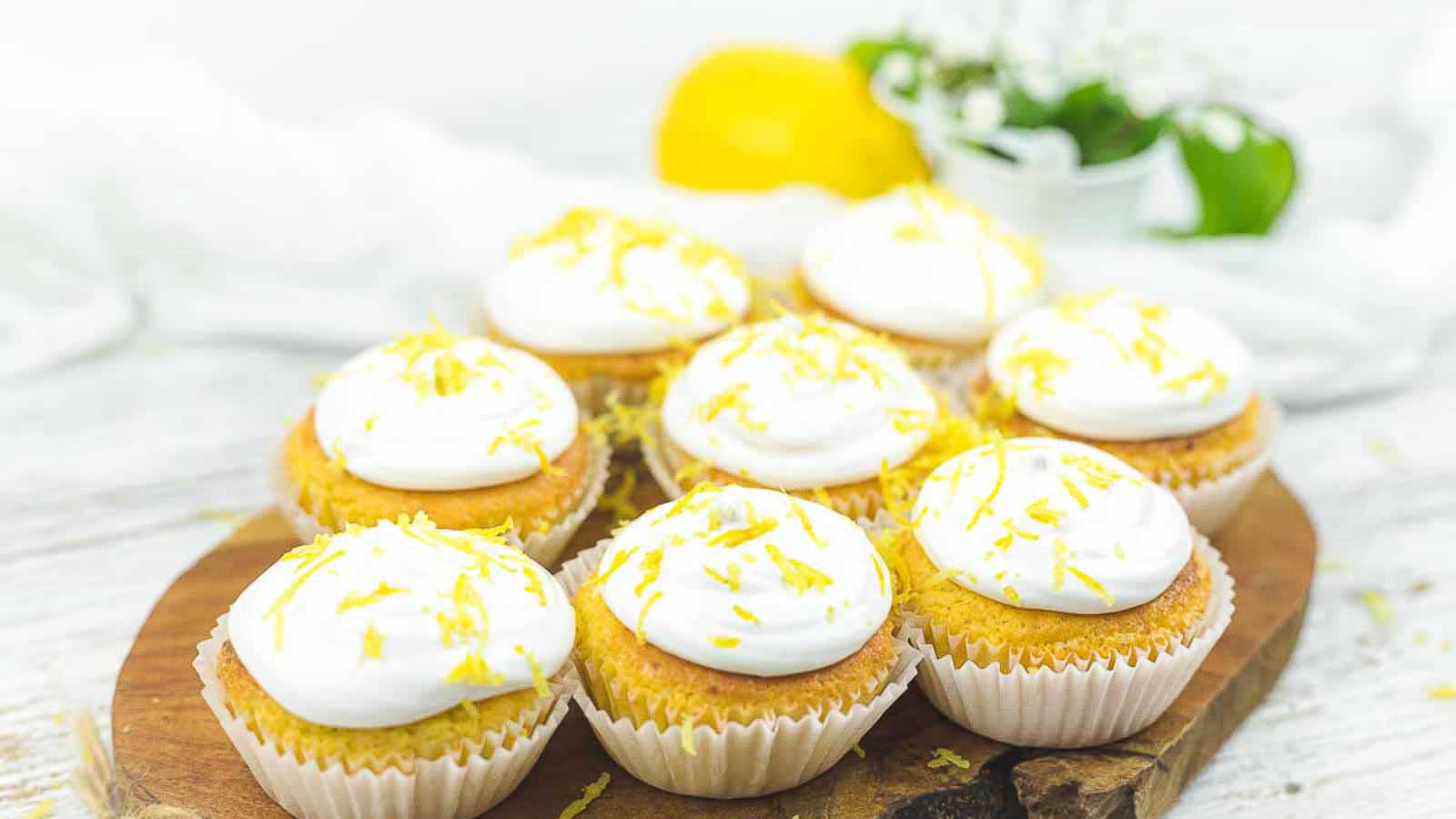 Nine lemon cupcakes with white frosting and lemon zest sit on a round wooden board. A lemon and some greenery are blurred in the background.