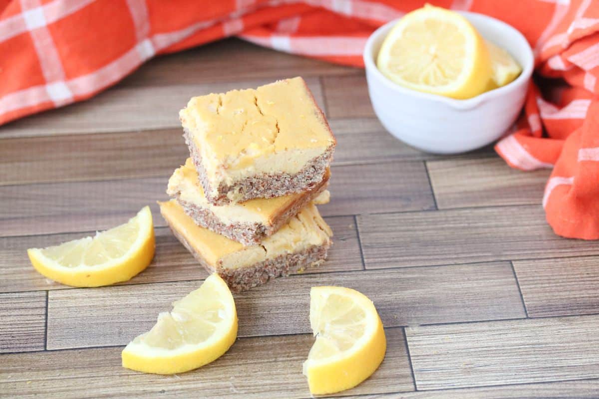 Three stacked lemon bars on a wooden surface with lemon wedges in front and a white bowl of lemon slices in the background. An orange checkered cloth is partially visible.