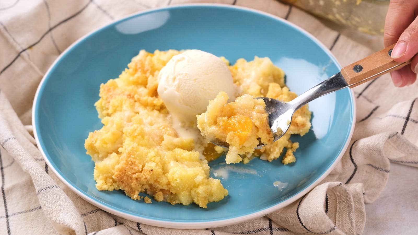 A serving of peach cobbler with a scoop of vanilla ice cream on a blue plate; a hand holds a spoonful of the dessert.