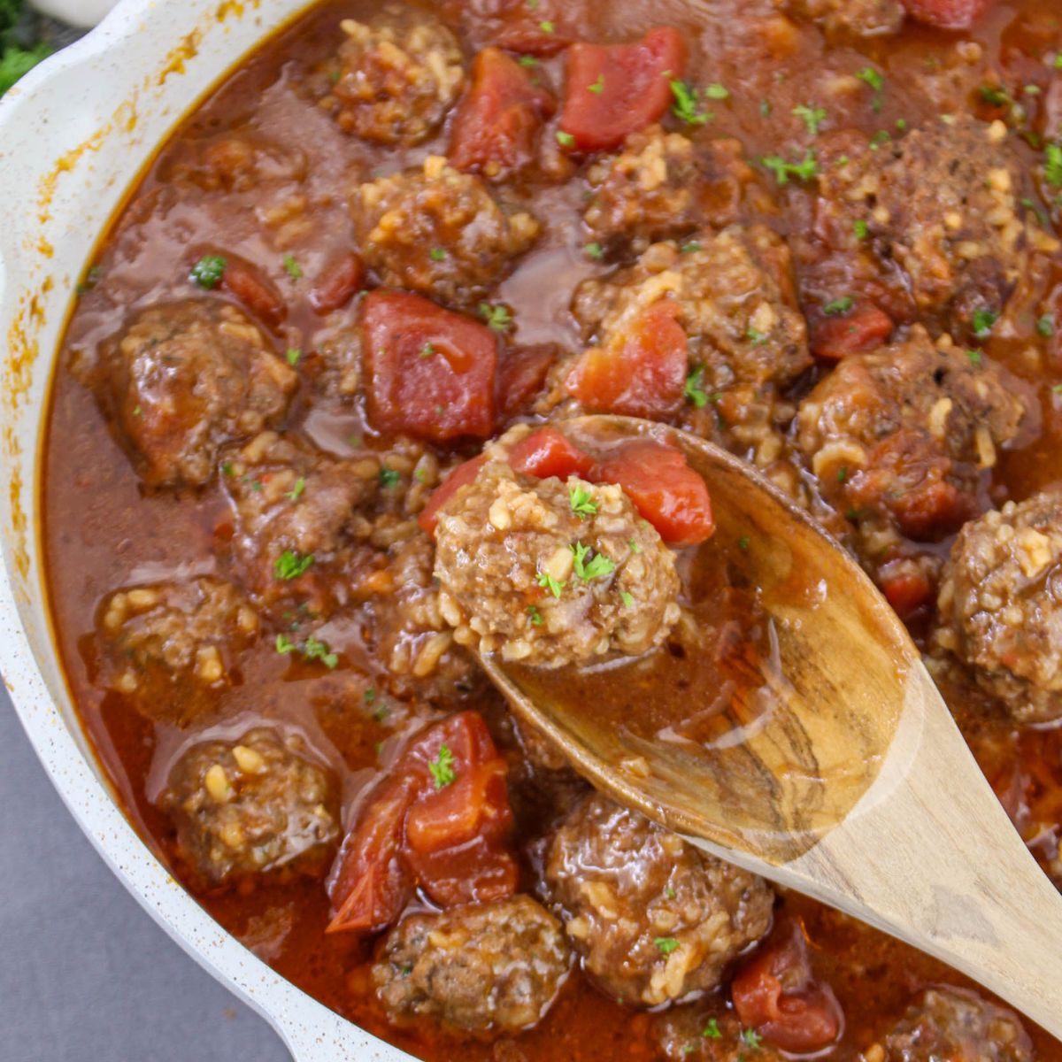 Porcupine Meatballs in tomato sauce with diced tomatoes and herbs simmer in a white pot, while a wooden spoon lifts one tender meatball.