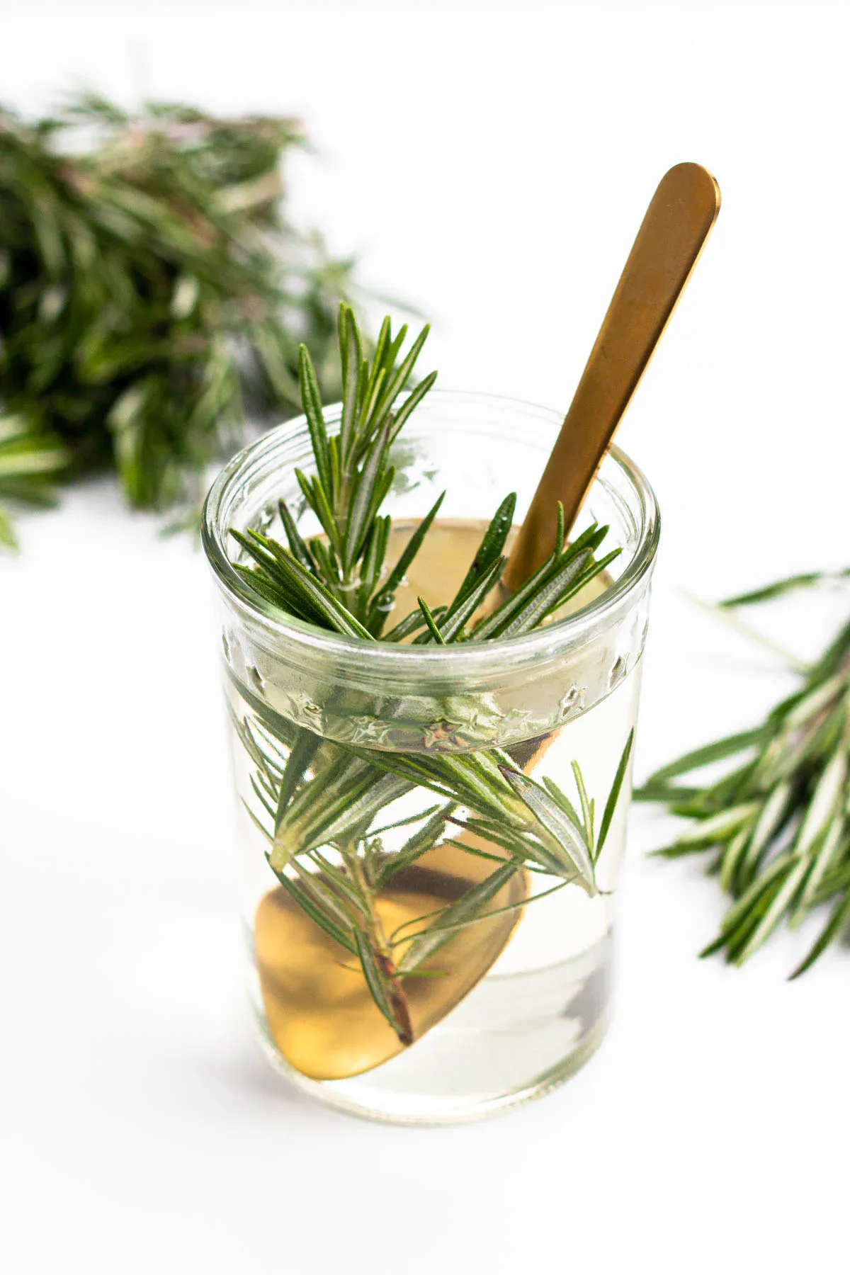 A glass filled with clear liquid, rosemary sprigs, and a gold spoon, with additional rosemary in the background on a white surface.