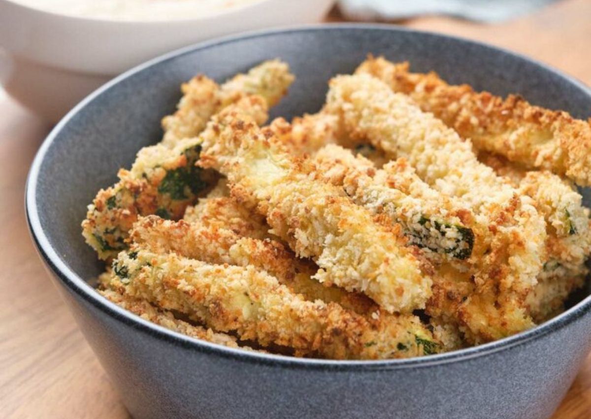 A bowl filled with crispy, breaded zucchini fries sits on a wooden surface, with a white bowl of dipping sauce in the background.