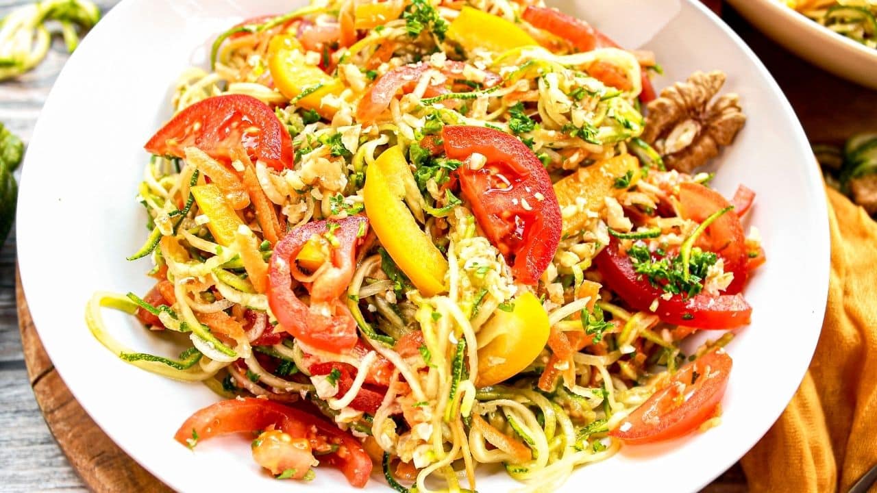 A plate of spiralized zucchini noodles mixed with sliced tomatoes, yellow bell peppers, herbs, and walnuts.