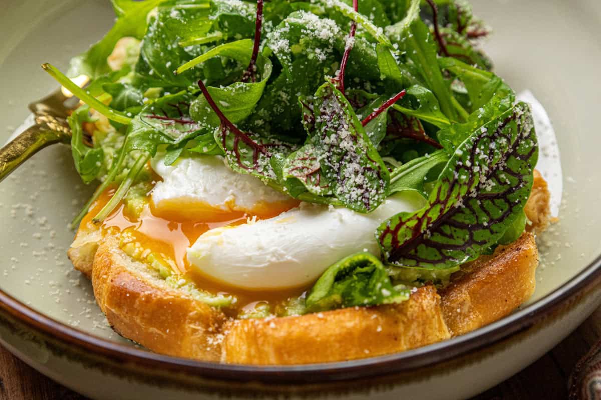 A poached egg on toast with greens, accompanied by creamy avocado and topped with grated cheese, served on a plate.