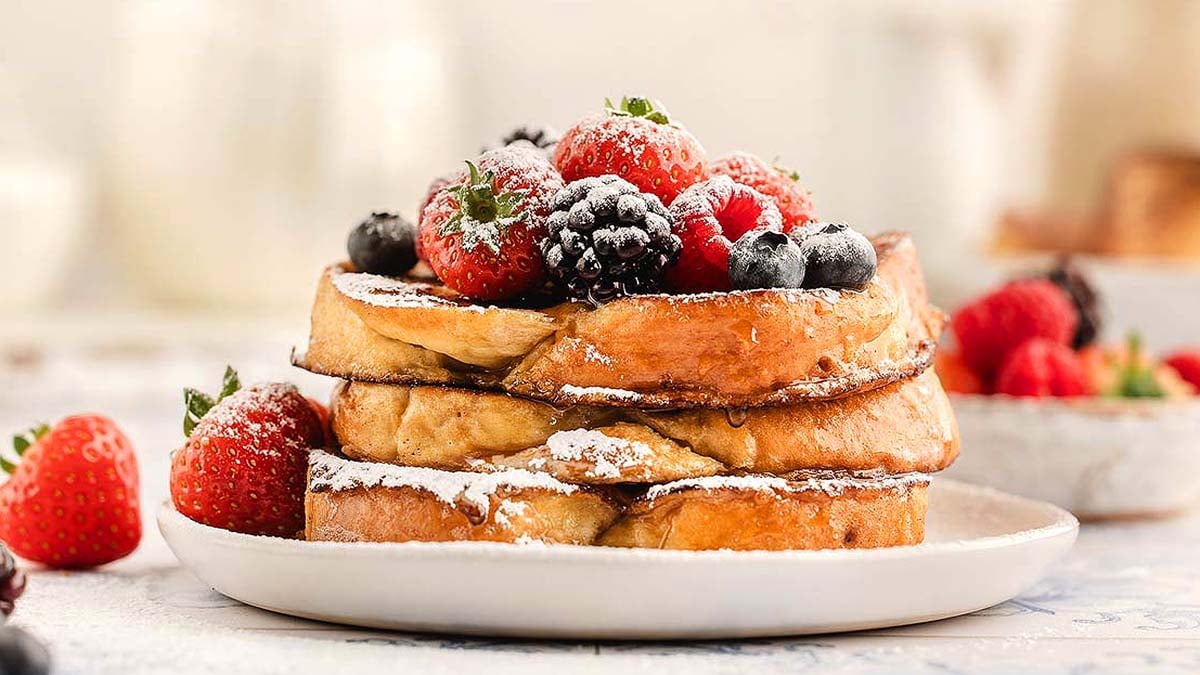 Three slices of French toast stacked on a plate, topped with powdered sugar, strawberries, blackberries, and blueberries.