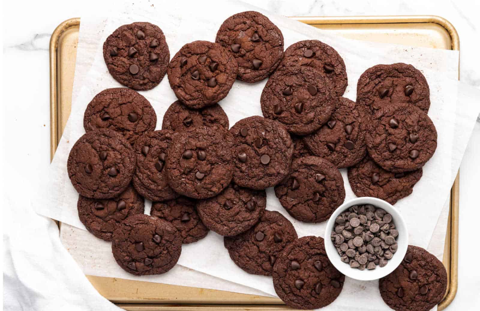 A baking tray with several chocolate cookies on parchment paper, next to a small bowl filled with chocolate chips.
