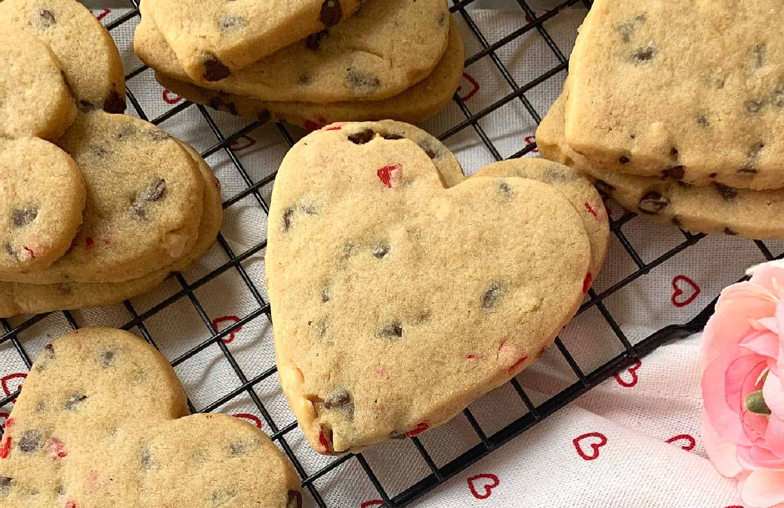Heart-shaped chocolate chip cookies with red candy bits are cooling on a black wire rack. A pink flower and heart-patterned cloth are partially visible.