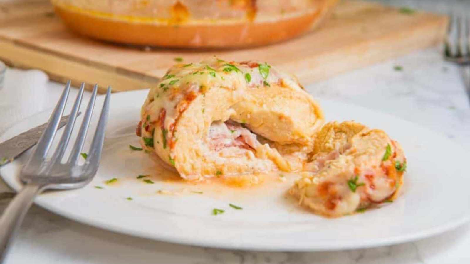 A stuffed chicken breast, topped with melted cheese and herbs, sits on a white plate with a fork, with a portion cut out to reveal the filling.