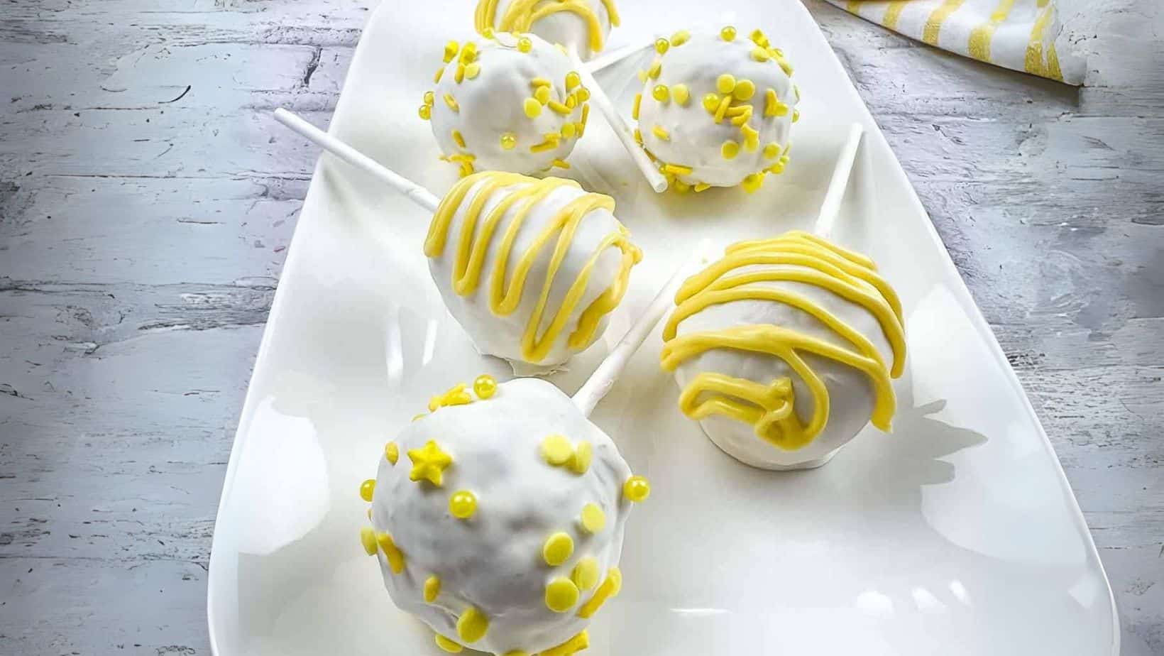 Five cake pops coated in white icing with yellow decorative toppings and drizzle, arranged on a white plate on a light wooden surface.