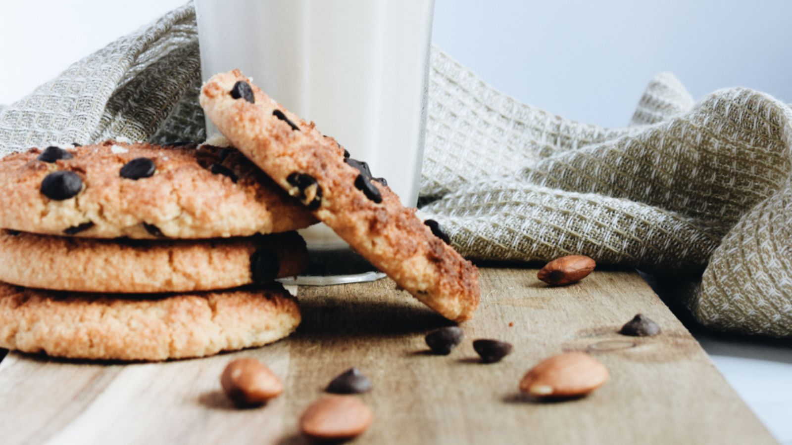 Three chocolate chip cookies are stacked on a wooden surface next to a glass of milk, with scattered almonds and a textured cloth in the background.