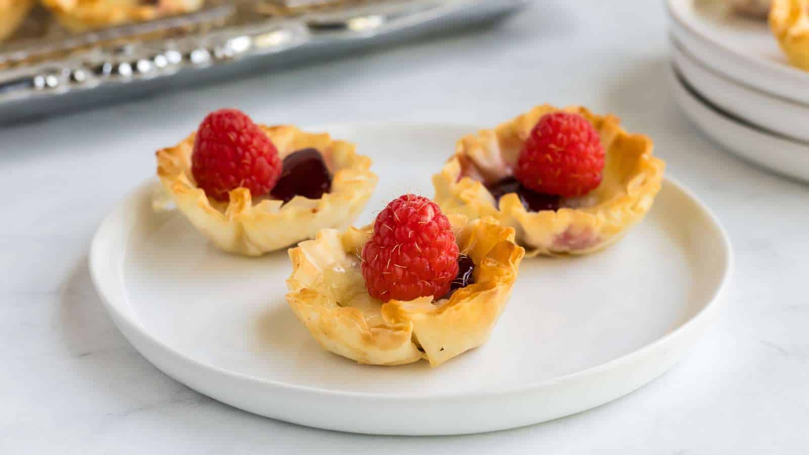 Three mini phyllo pastry cups filled with jam or custard and topped with a fresh raspberry, arranged on a white plate.
