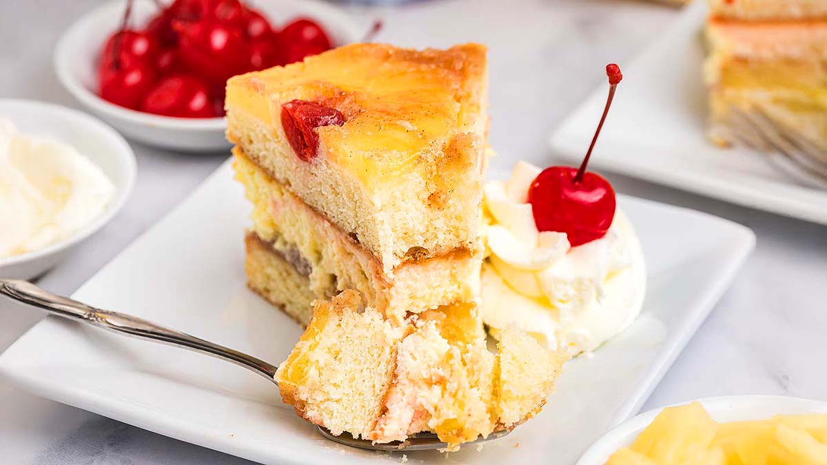 A slice of layered cake with fruit filling, topped with a cherry, served on a white plate with whipped cream and a spoon; a bowl of cherries is in the background.