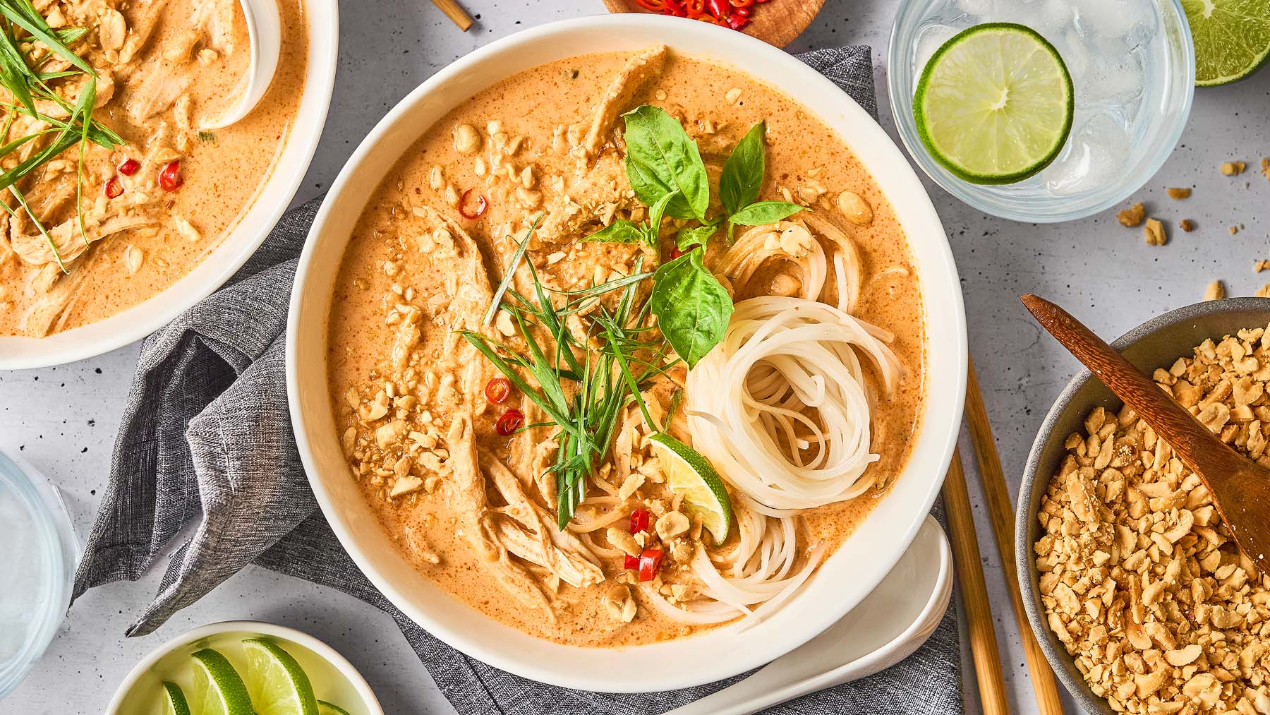 A bowl of peanut noodle soup garnished with herbs, sliced chili, crushed peanuts, and lime, surrounded by lime wedges, a bowl of chopped peanuts, and a glass of water with lime.